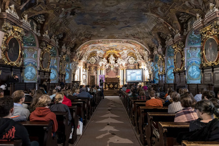 Menschen in einem prunkvollen, ausgemalden und mit goldenen Ornamenten verziertem Gewölbe von hinten zu sehen, weit hinten steht ein Redner.