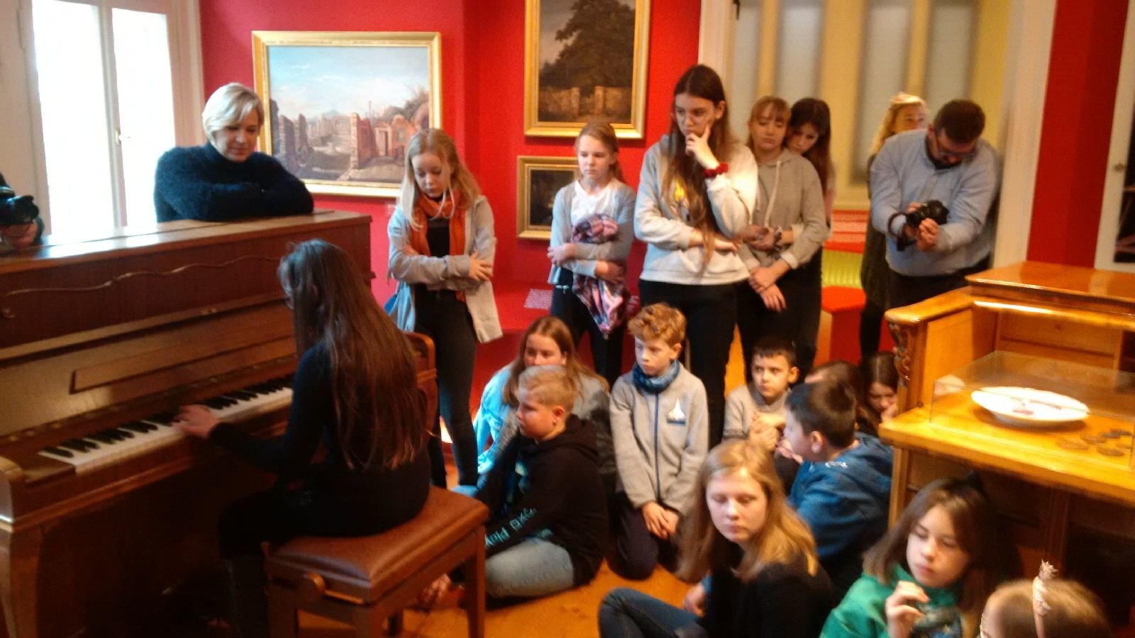 Mädchen mit langem braunem Haar spielt Klavier, Erwachsene und Kinder stehen um sie herum oder sitzen auf dem Fußboden. An den roten Wänden hängen Bilder im Goldrahmen. 