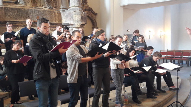 Männer und Frauen haben Partituren in der Hand und singen im Chor. Einige sitzen, andere stehen. Im Hintergrund des Raumes ein historisches Relief.